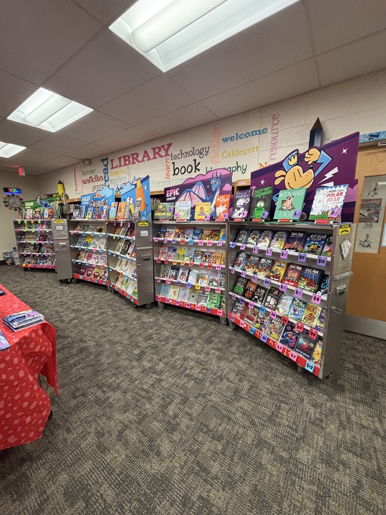 A library with rows of bookshelves filled with colorful books. A table with a red tablecloth is on the left.