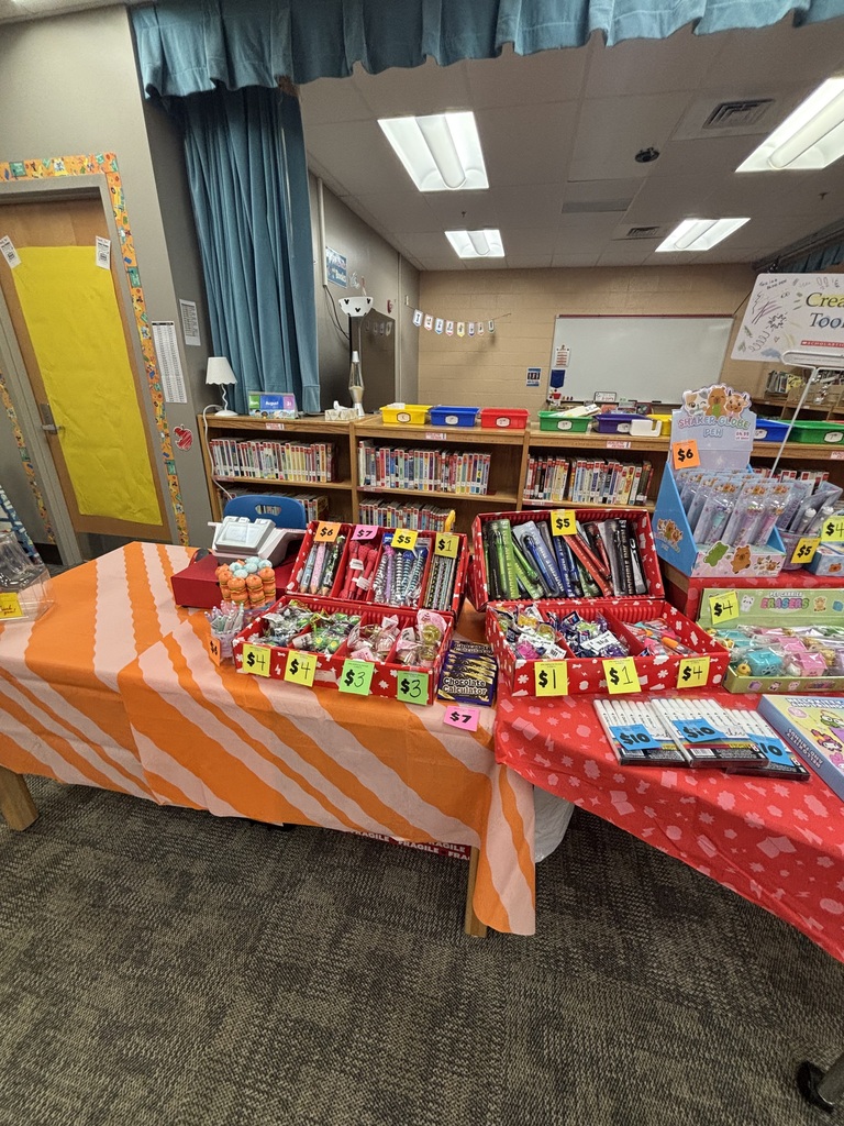 Two tables in a classroom. One has orange and white striped tablecloth with books and stationery. Other table has red and white tablecloth.