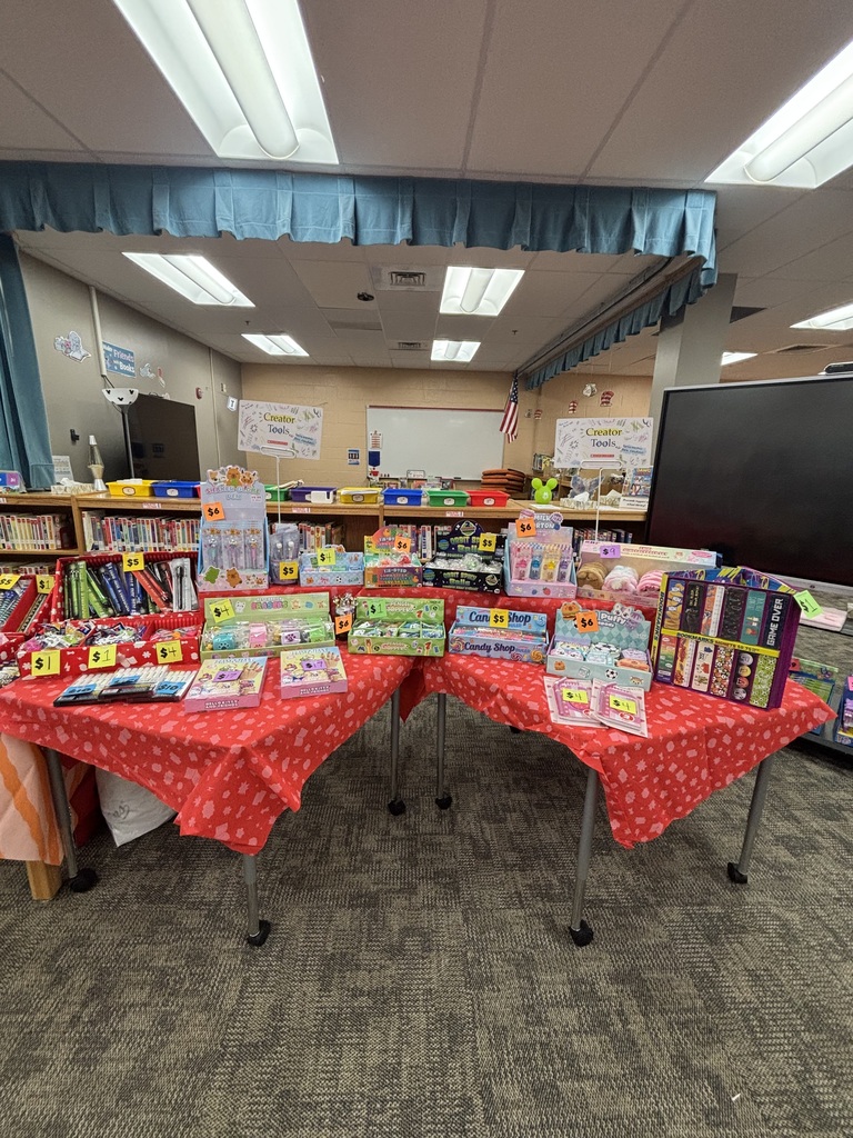 Library area with two tables covered in red fabric, each displaying various books and toys. Background features bookshelves and ceiling lights.