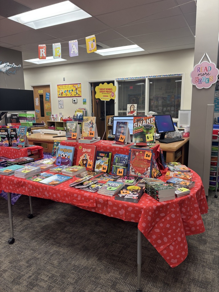 A library has a long table covered with books. A monitor and keyboard are on a wooden desk.