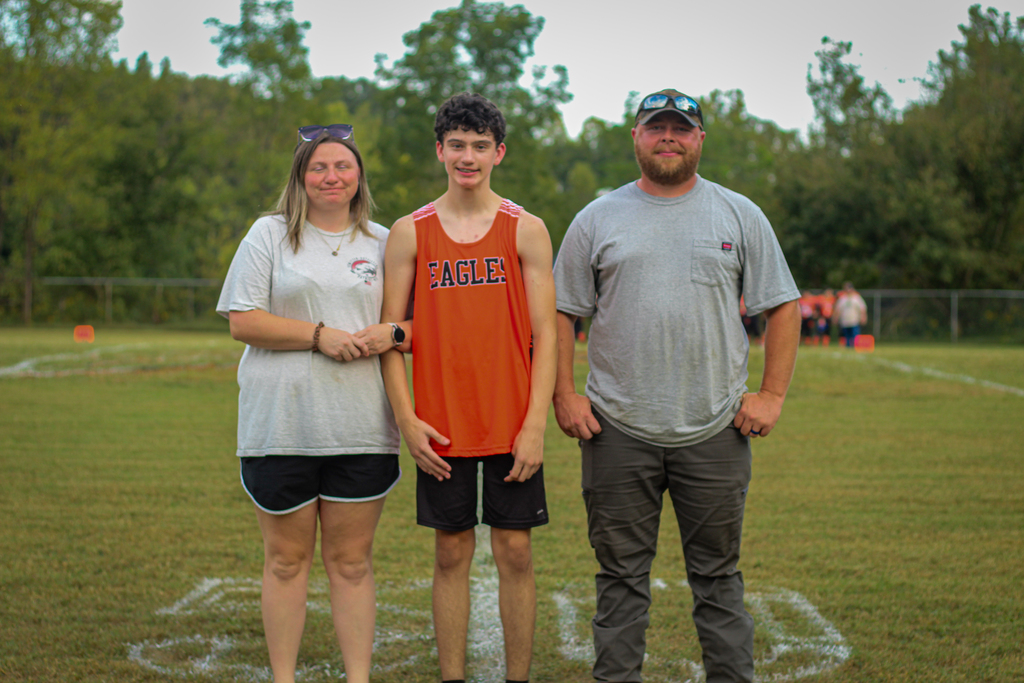 Three people stand on a grassy field. A woman, a boy in an orange shirt, and a man in a gray shirt.