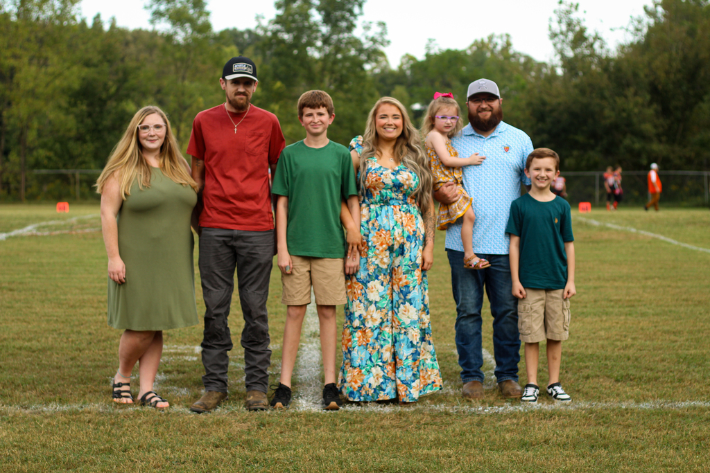 Six people on a grassy field, wearing casual clothing. A woman carries a child on her hip.