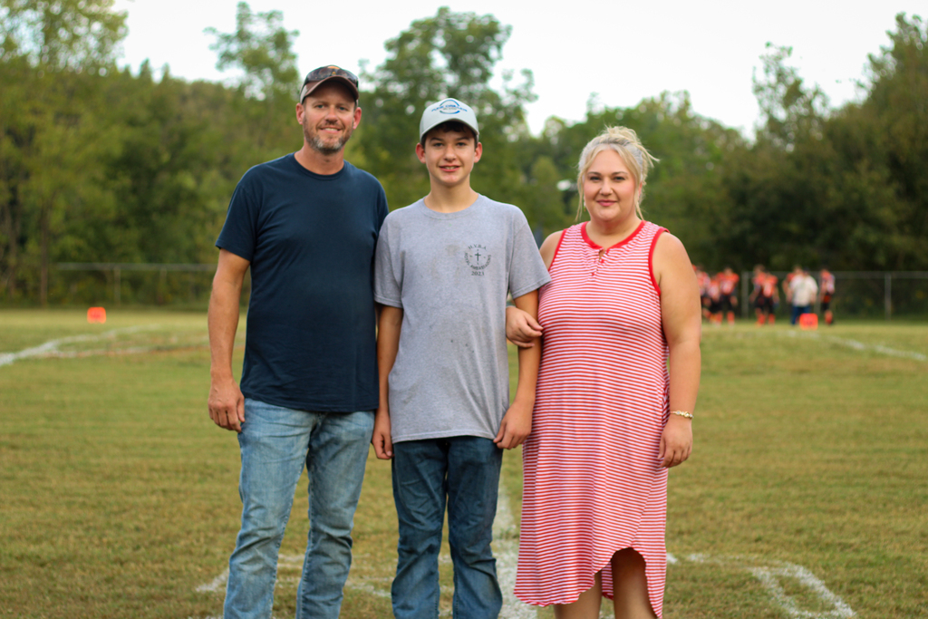 Three people on a football field, two men and one woman, with trees in the background.