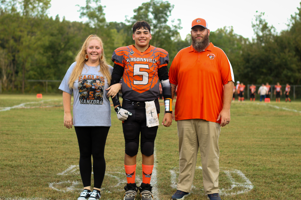 Three people stand on a field, with a football player in uniform between a man and a woman.