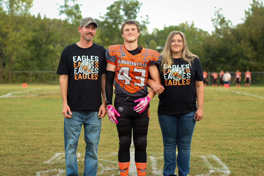 Three people stand on a field. A man and two women wear black shirts. The man wears a cap.