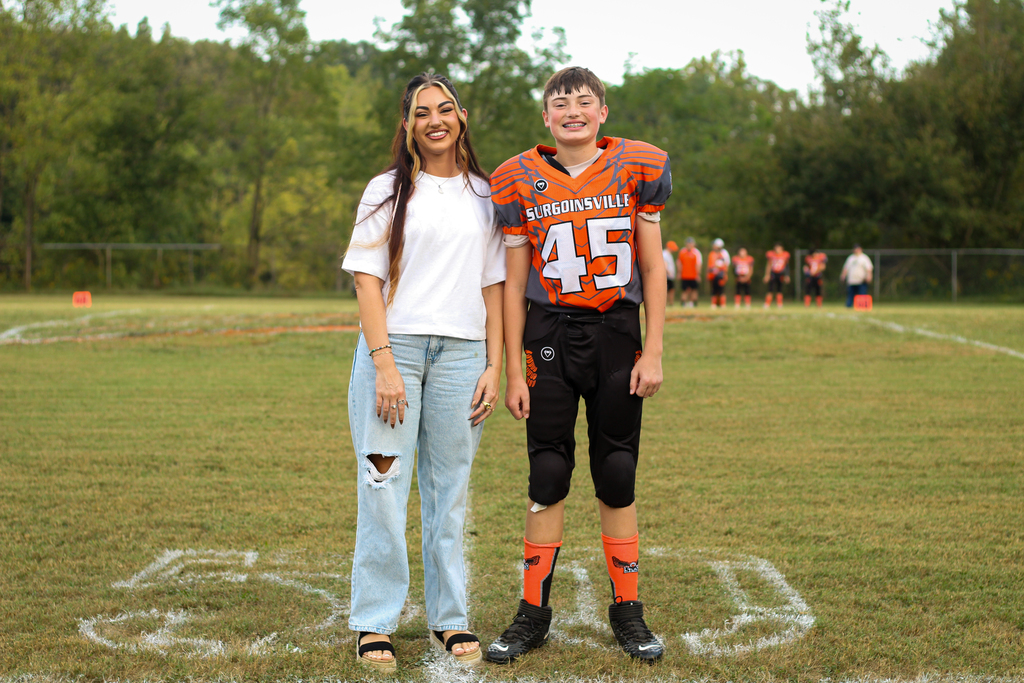 Two people on a field. A woman wears a white shirt and jeans; a boy has an orange football jersey.