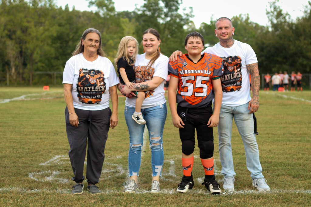 Four individuals in matching white T-shirts stand on a football field. A man in football gear stands between a woman and a child.