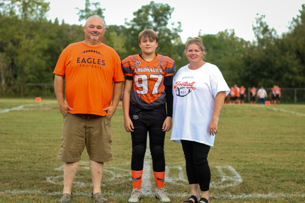 Three people stand on a football field. A man wears an orange shirt, a youth in sports gear, and a woman in white.