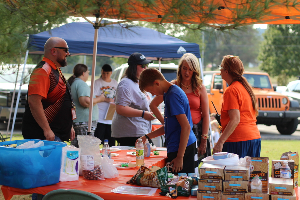 A group gathers around a table with food and supplies. An orange canopy provides shade.