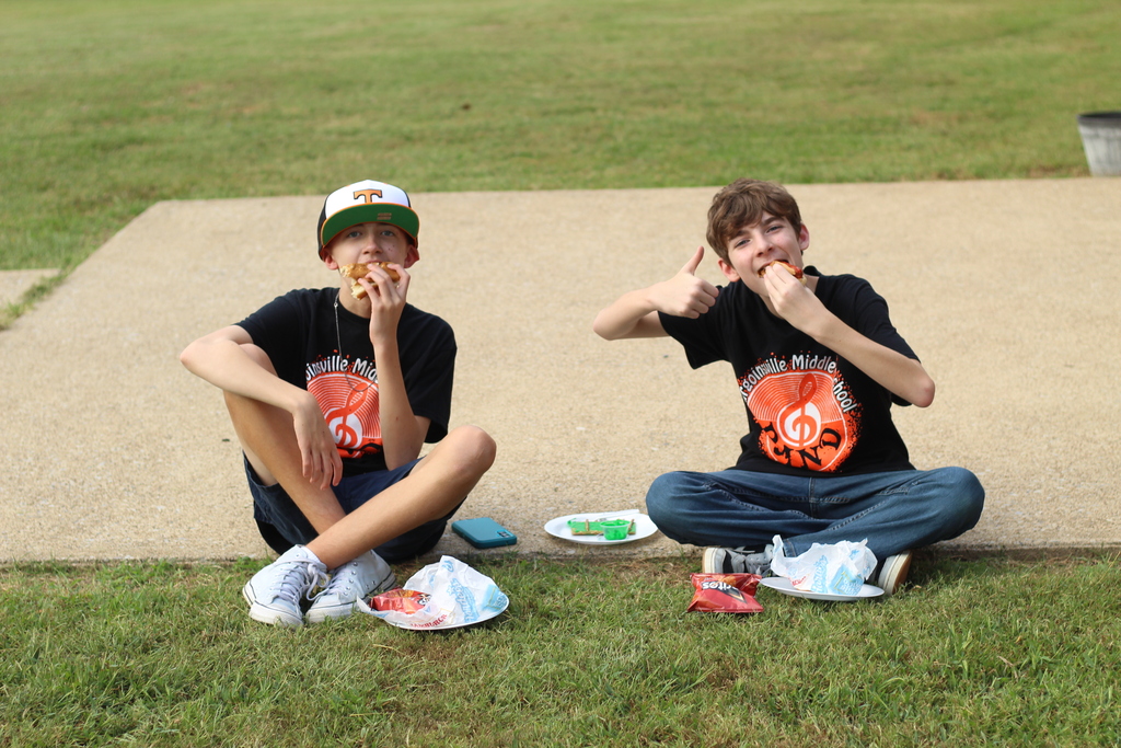 Two boys sit on grass, each eating hot dogs. One boy wears a cap and sneakers, the other wears a shirt with graphics.