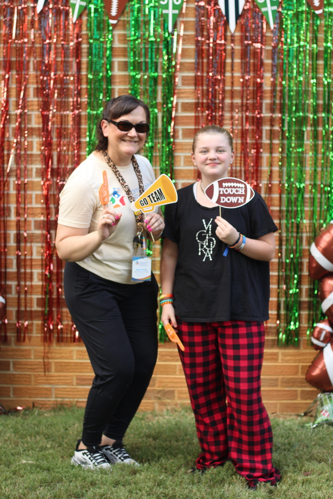Two people stand in front of a brick wall with festive decorations; one holds a sign, the other wears a football shirt.