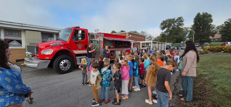 Children in front of a fire truck with two adults. The fire truck is parked on the street.