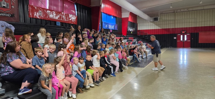 Group of children seated on bleachers, watching a man standing in the middle of the room.
