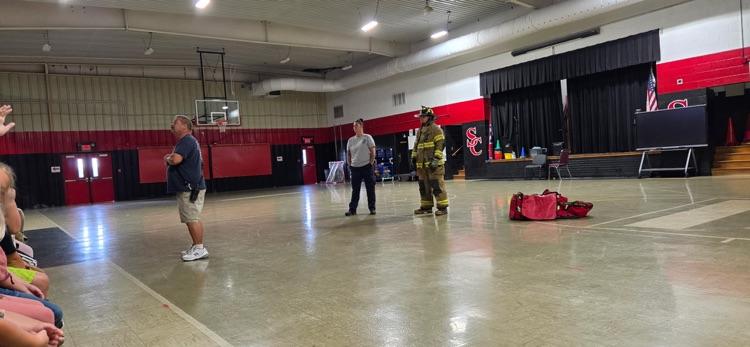 A firefighter stands in a gymnasium with a fire extinguisher. Two men in white shirts stand nearby.