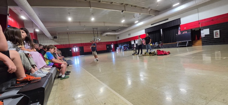 Indoor basketball court with red and white walls, bleachers with seated children, two people standing near the middle, and a person walking in the distance.