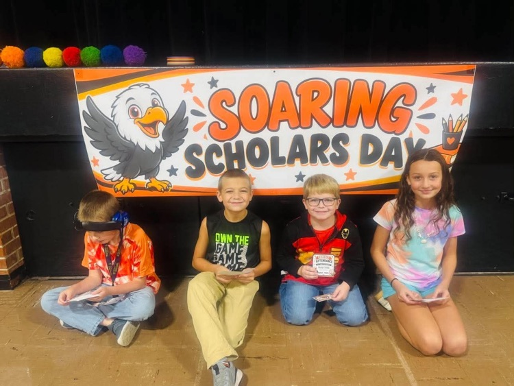 Four children sit in front of a banner that says "Soaring Scholars Day" with an eagle.
