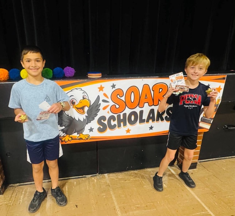 Two boys stand before a banner with an eagle and text reading "SOAR SCHOLARS". They hold papers and small objects.