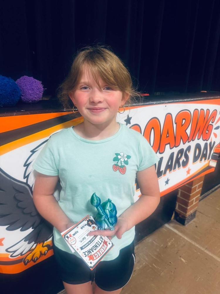 Girl with blonde hair stands with award, smiling at camera. Behind, banner with eagle and words.