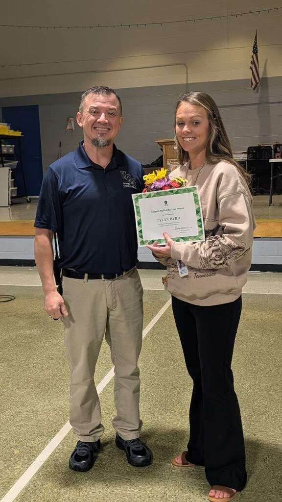 A man and woman stand together in a gym, both smiling, holding a certificate with flowers.