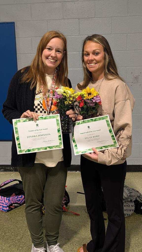 Two women smiling and holding certificates and flowers in a school hallway. A bag is on the floor behind them.