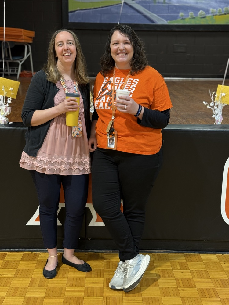 Two adult women stand next to each other holding drinks, one in yellow and one in white, in front of a stage.