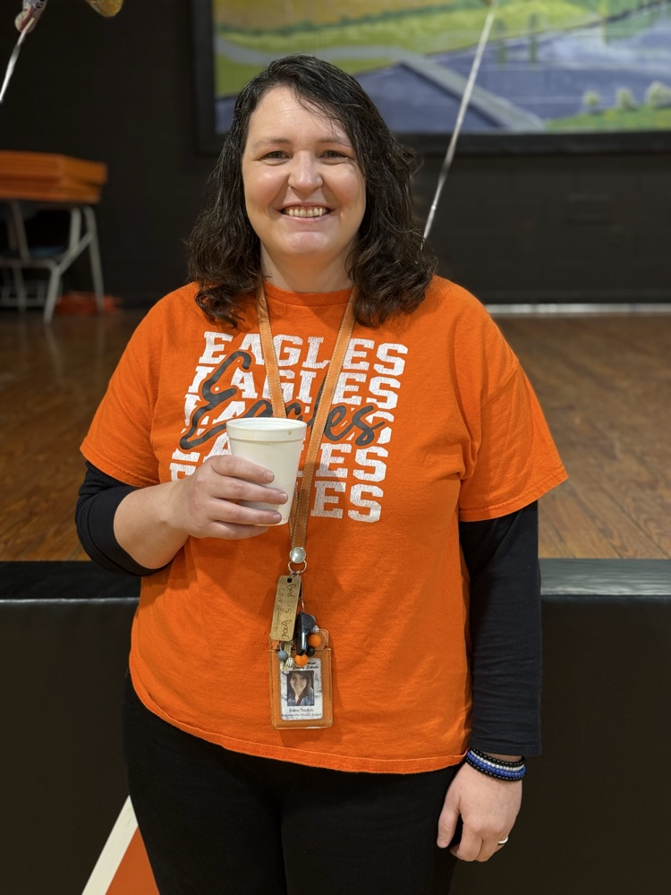 Woman in orange Eagles T-shirt holding a cup, standing in front of a stage in a gym.