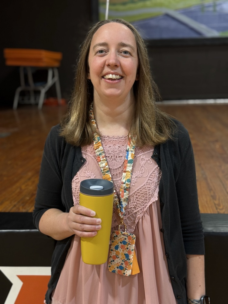 A smiling woman holds a yellow tumbler while standing against a black wall with an orange star.