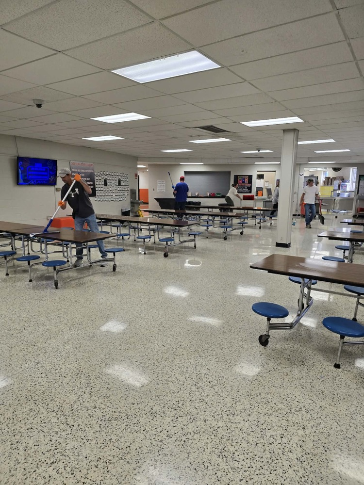 A large room with white tile floor, tables and chairs. A person is sweeping the floor.