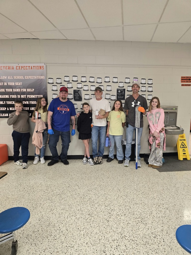 Several students and adults stand in a hallway. They wear gloves, some hold brooms, and a sign reads "cafeteria expectations" on the wall.