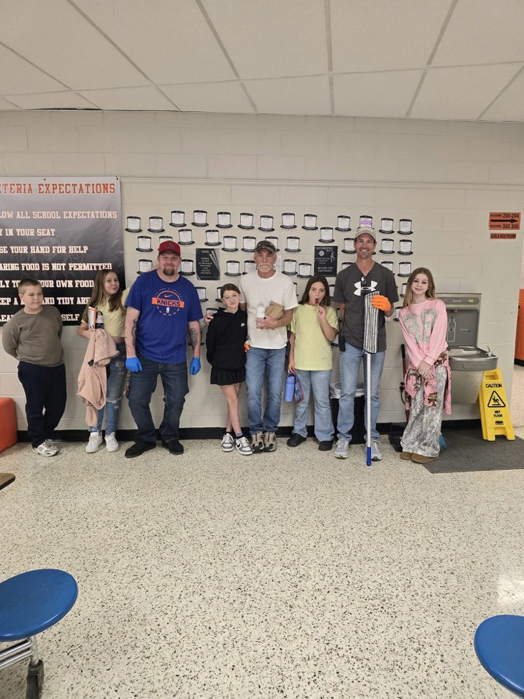 A group of adults and children pose with cleaning supplies in a hallway with white walls.