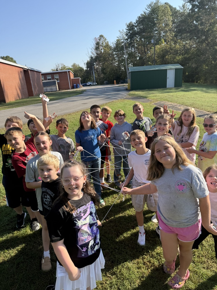Group of children stand on grass holding strings with hands raised, in front of school buildings.