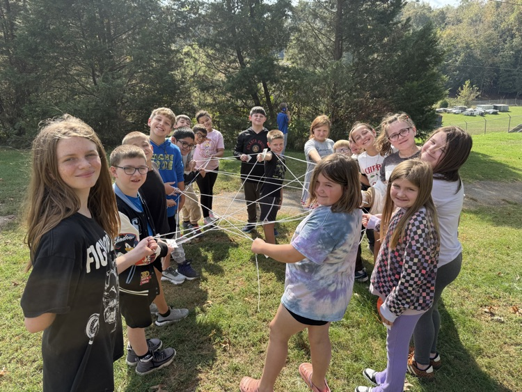 Students gather outdoors for an activity with strings and sticks; trees and a road visible in the background.