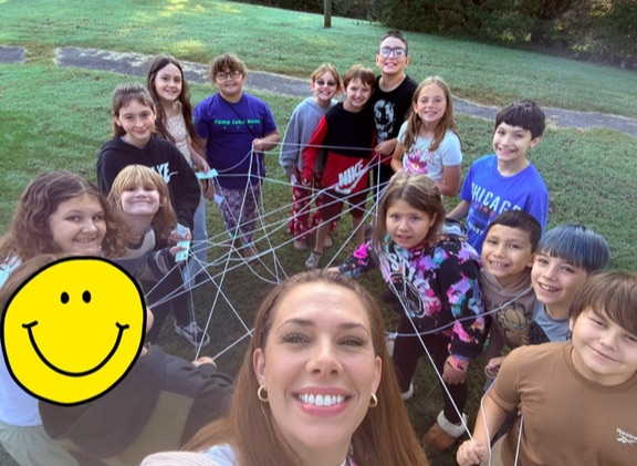 Children and a smiling adult stand in a grassy area, holding a large web. A yellow smiley face sticker is in the top left corner.