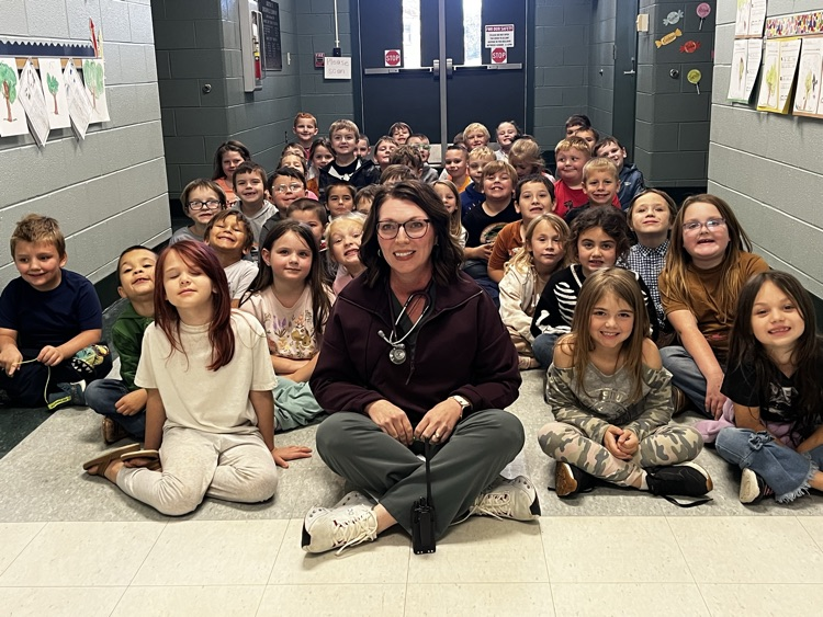 A teacher and students sitting on the floor, some smiling, inside a school hallway.