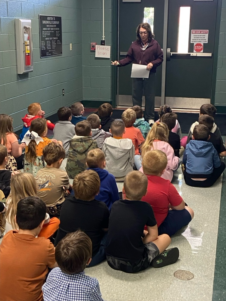 An adult woman stands in a hallway, reading from a paper to a circle of seated children.