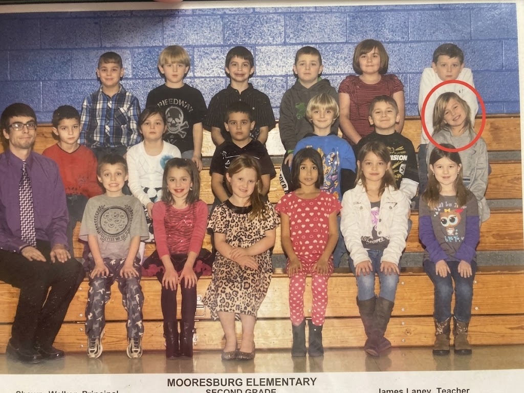Teacher with class; children in rows on bleachers, most wearing winter clothing; one with red circle around face.
