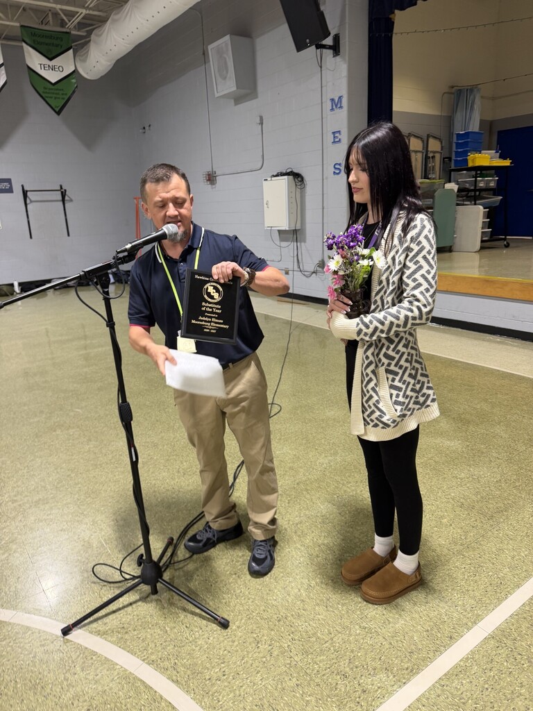 Man standing at a microphone holding paper, speaking next to a woman holding flowers.
