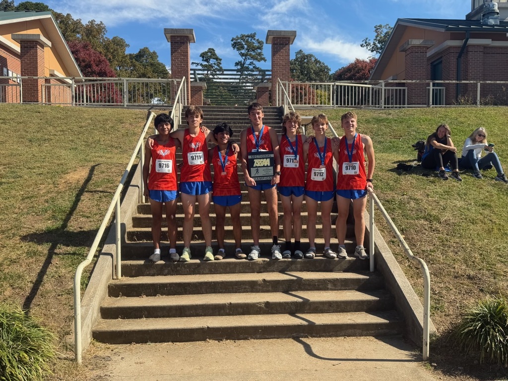 Team members in uniforms stand on outdoor stairs, holding medals, with a grassy hill and buildings in the background.