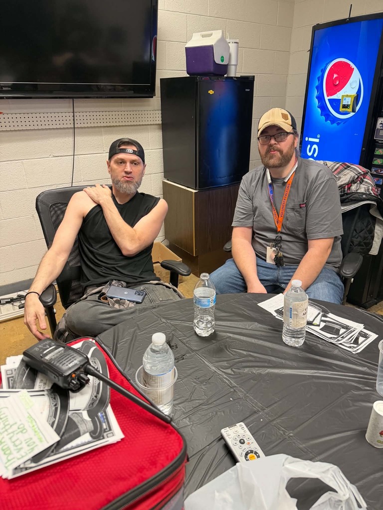 Two men in a small room. One sits in a chair with crossed arms. Another sits at a table with bottles of water and remote controls.