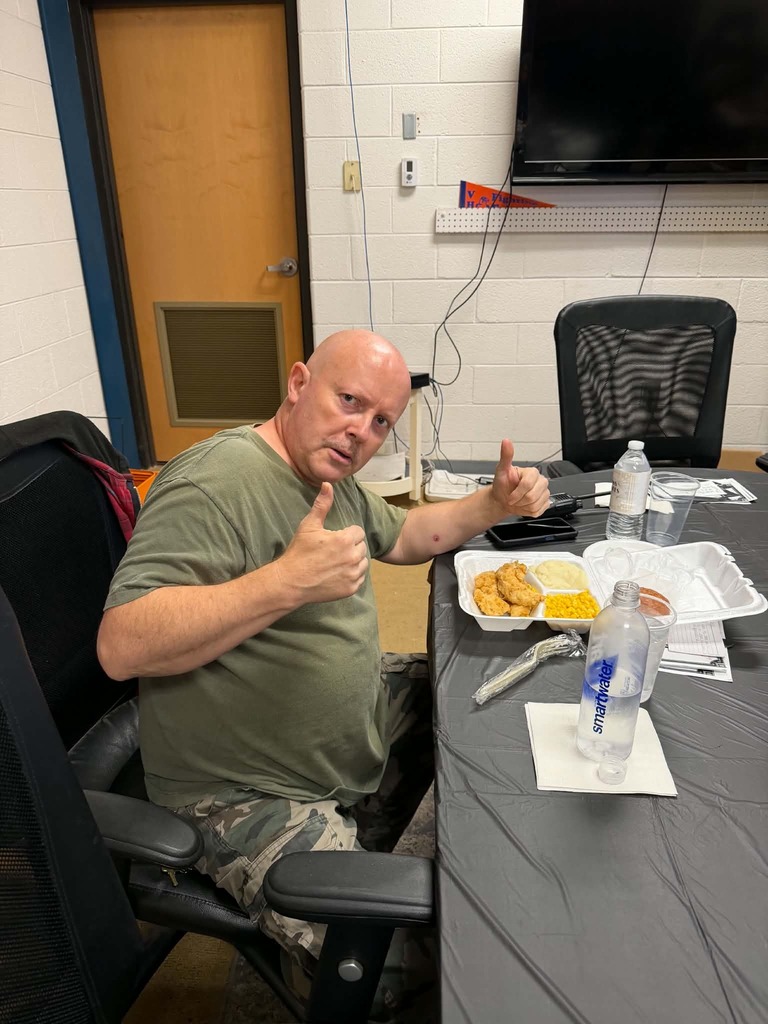 Person giving a thumbs-up while sitting at a table with food, water, and other items.