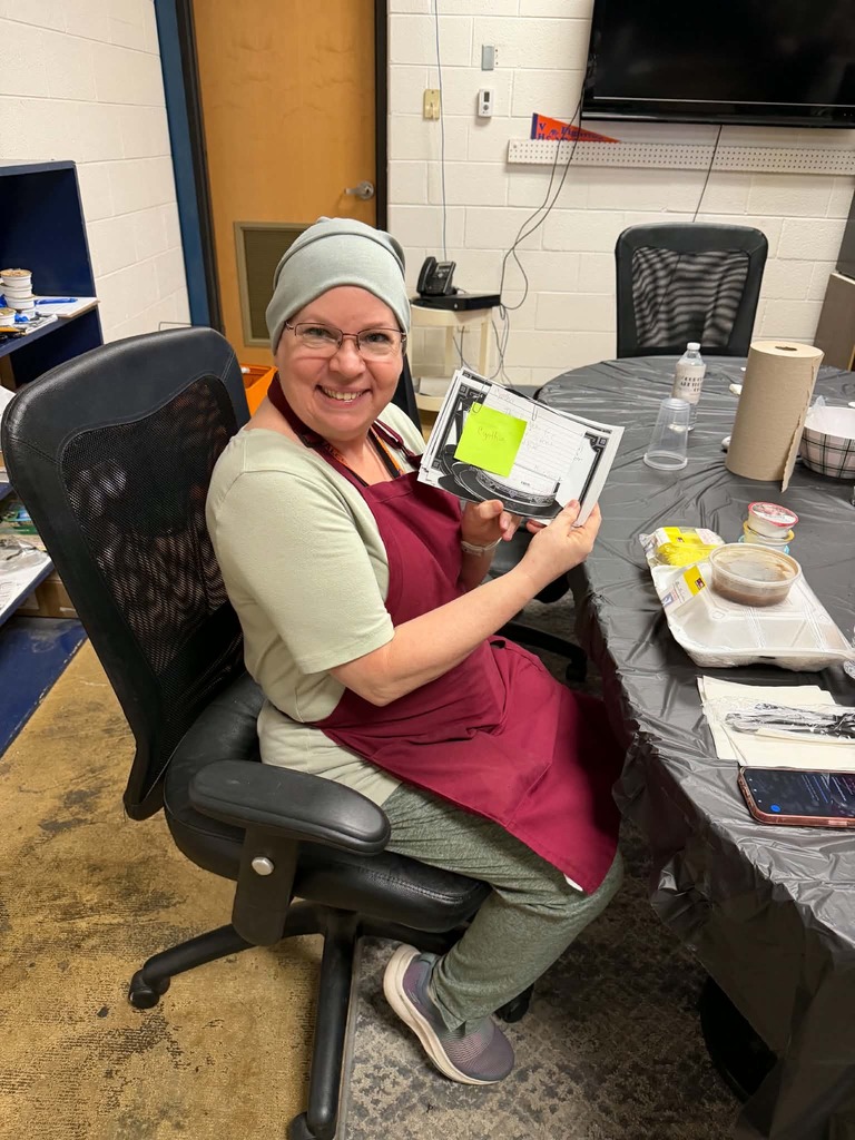 Woman in an apron seated at a table, holding a book and smiling for a photo.