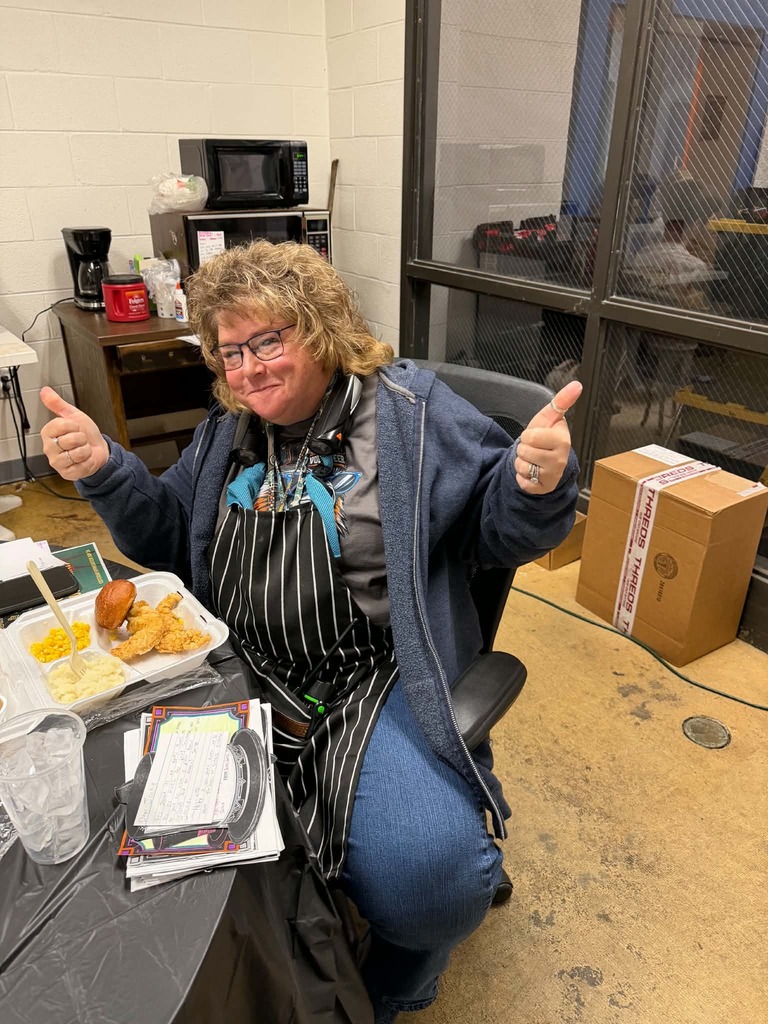 A woman with short hair wearing glasses sits at a table giving two thumbs up.