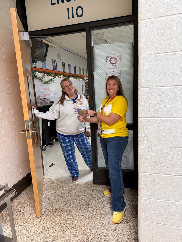 Two women stand at a classroom door, one holding a small object, the other holding a door handle.