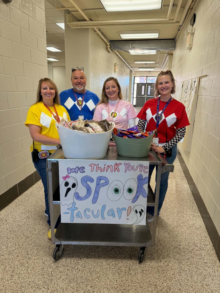  people in costumes stand near a table with buckets of candy. Behind them, a hallway with tiled floor and bright lights.