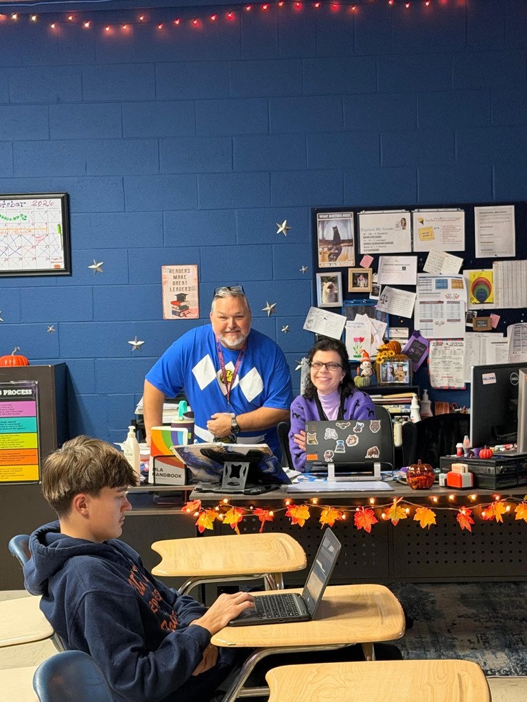 Three people in a room, two adults and a child, seated at desks. Blue wall with bulletin board, orange fall leaves.