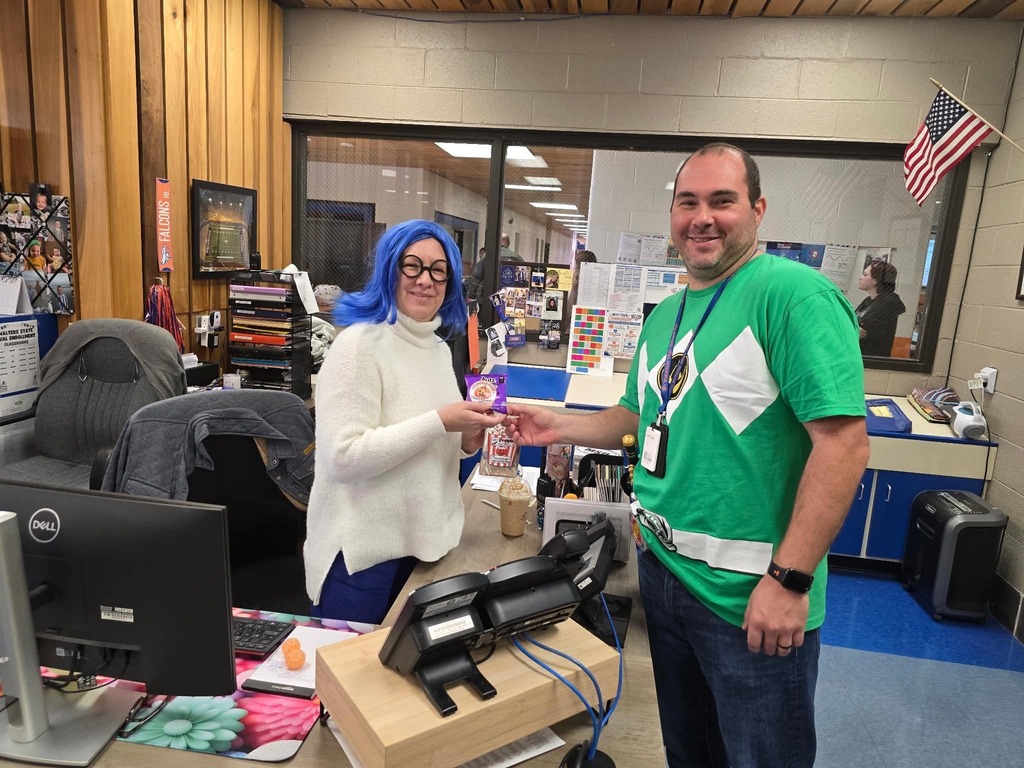 Person with blue hair stands behind desk with a person in a green shirt.