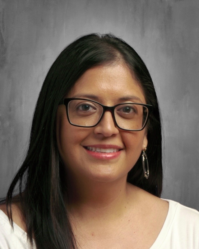 A smiling woman with long black hair, glasses and white shirt poses against a gray background.