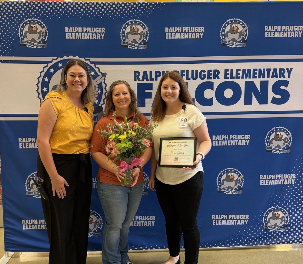 Three women standing next to each other with the woman in the middle carrying a vase with flowers and the woman on the right carrying a certificate for volunteer of the year.