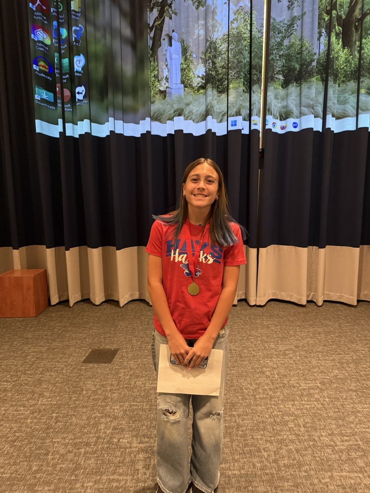 A female Barton Middle School student with shoulder brown hair, wearing a red Hays Hawks t-shirt with a medal around her neck, smiling at the camera.