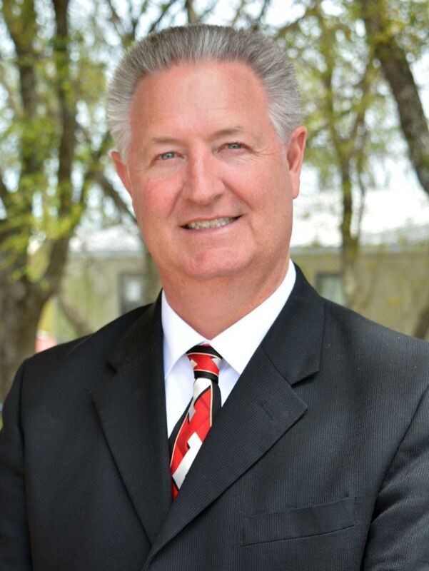 An man in a suit and tie smiling for a photo outdoors with trees in the background.